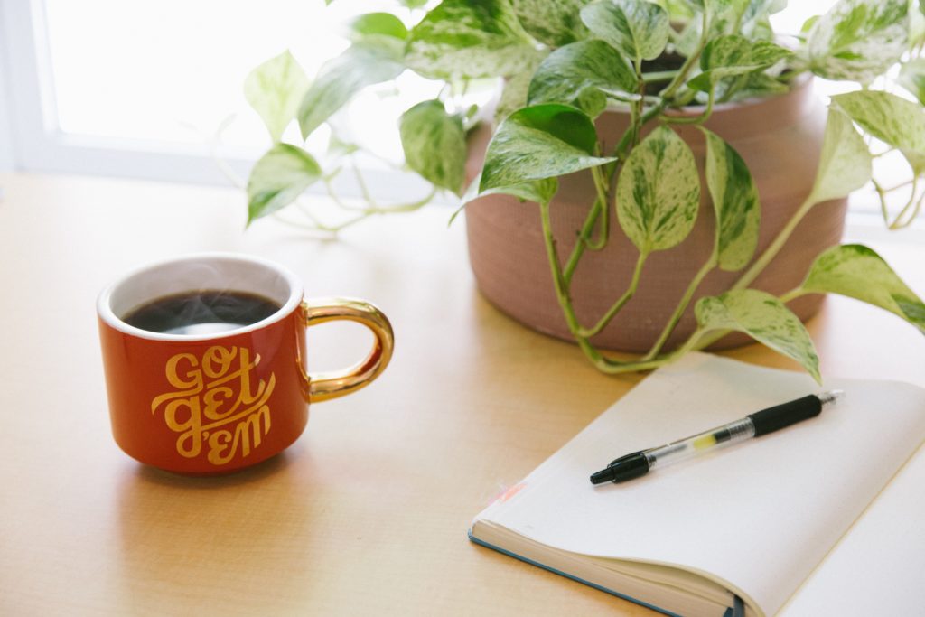 red coffee mug that says "go get em" next to a notebook, pen, and plant on a desk